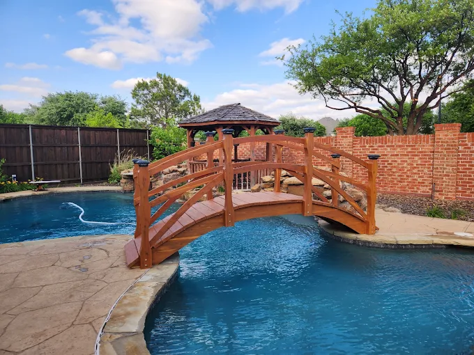 Handcrafted cedar high-rise pool bridge with curved rails and solar post lights spanning a backyard swimming pool, built by Redwood Bridges.