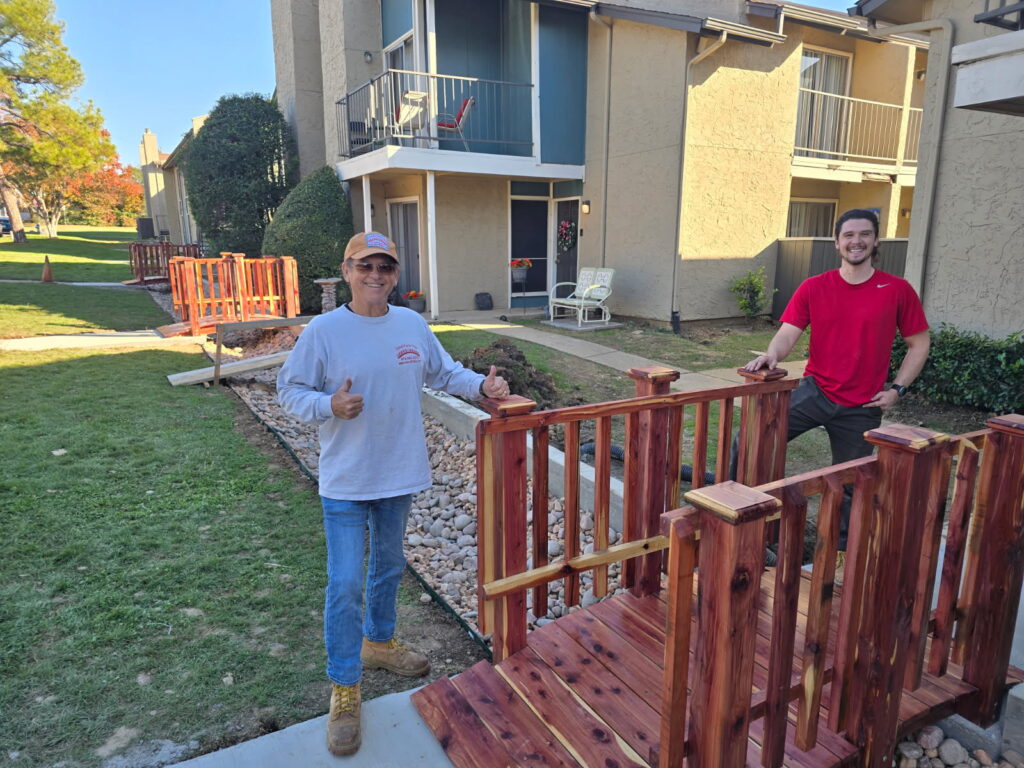 Two installers standing beside a newly installed cedar garden bridge at Holly Springs Apartments, showcasing completed footbridge project.