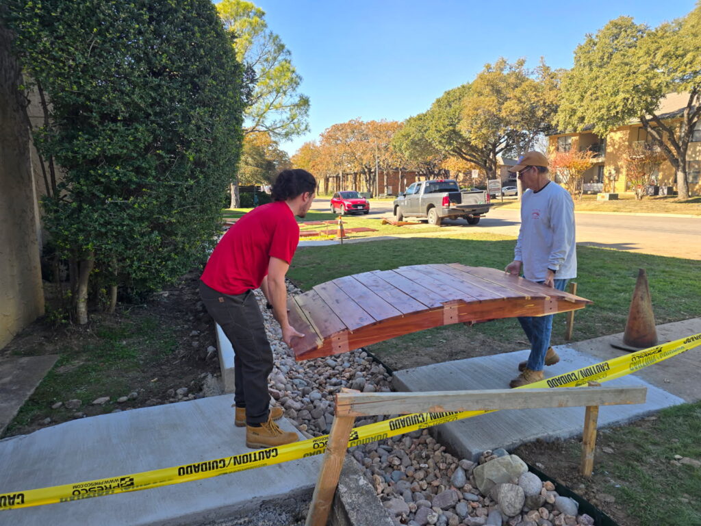 Installation of a cedar garden bridge at Holly Springs Apartments, with two workers placing the bridge over a concrete path and rock drainage.