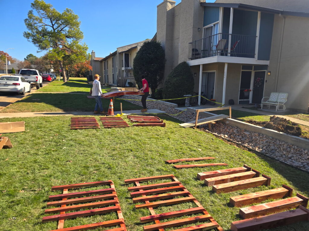 Cedar garden bridge components laid out on grass during installation at Holly Springs Apartments, with workers preparing for assembly.