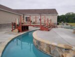 A custom 16ft handcrafted cedar step garden bridge with rope railings spanning over a backyard concrete lazy river swimming pool next to a patio.