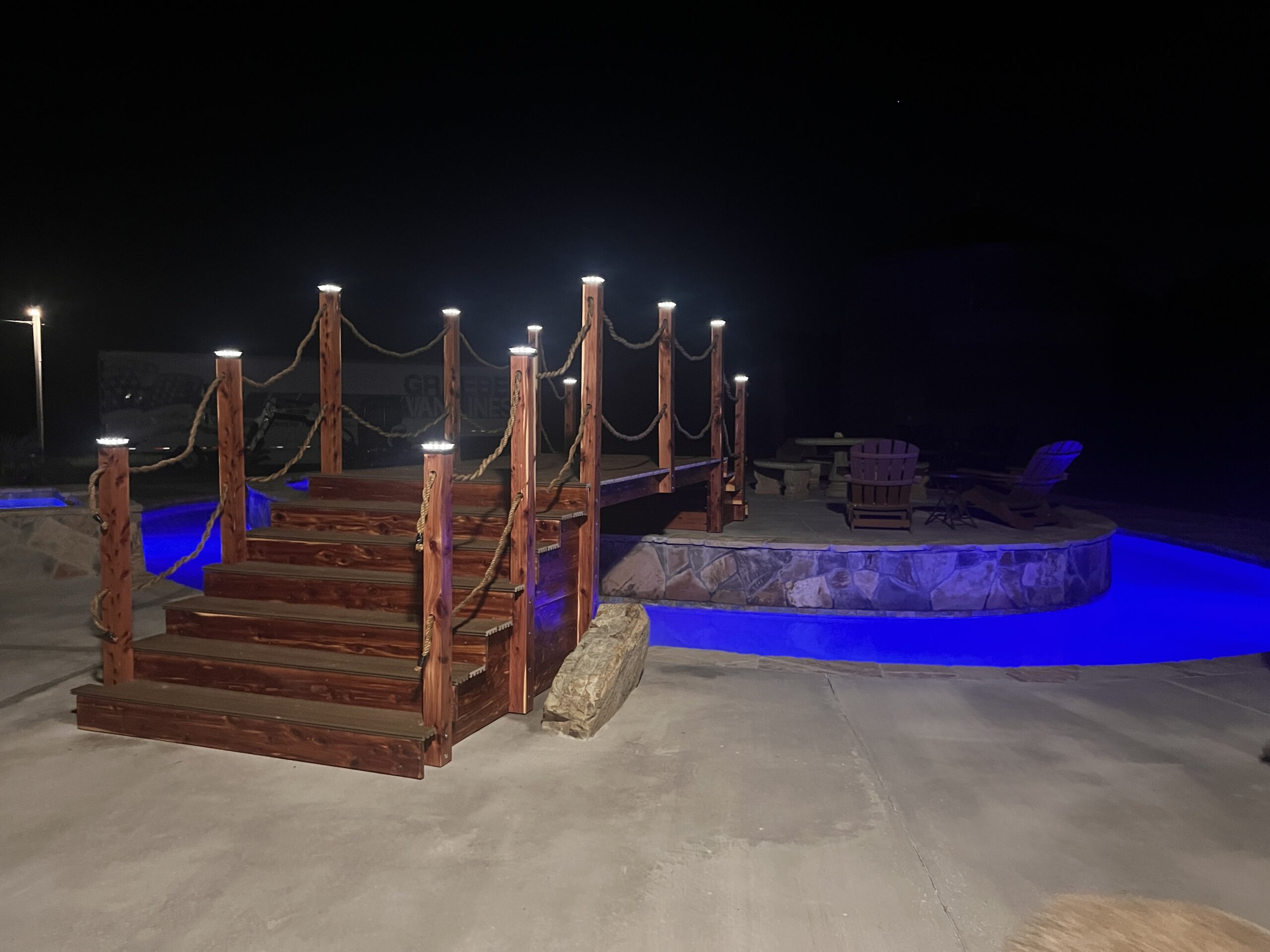 A custom 16ft handcrafted cedar step garden bridge with rope railings and illuminated post cap lights spanning a glowing blue lazy river pool at night.