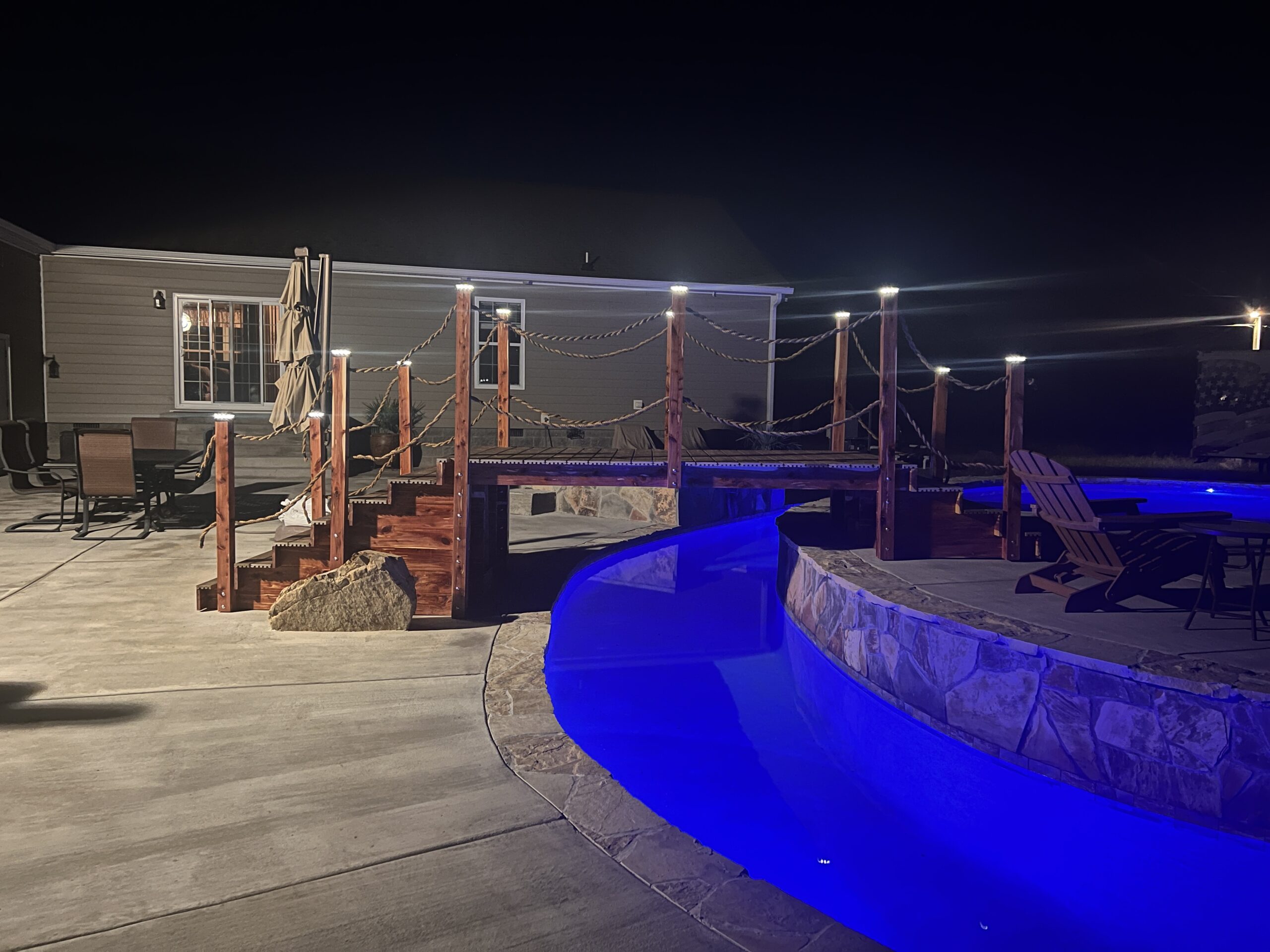 A custom 16ft handcrafted cedar step garden bridge with rope railings and illuminated post cap lights spanning a glowing blue lazy river pool at night.