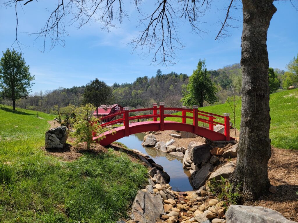 A 20-foot vibrant red Japanese-style high arch wooden footbridge built over a rocky stream on a landscaped property, handcrafted by Redwood Bridges.