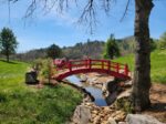 A 20-foot vibrant red Japanese-style high arch wooden footbridge built over a rocky stream on a landscaped property, handcrafted by Redwood Bridges. high arch garden bridge